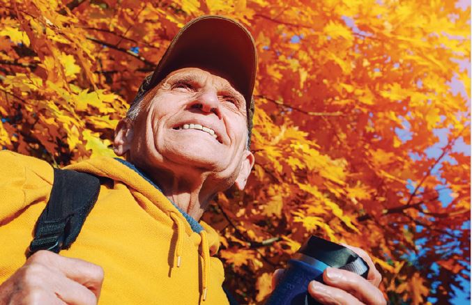 Photo of older person with hat on standing below a tree with its yellow autumn leaves