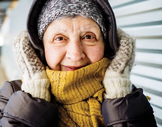 Older lady wearing mittens, a beanie and a warm jumper and jacket with hood.