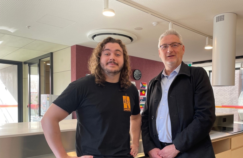 Jonas stands with Anglicare's General Manager Housing and Community Services Noel Mundy in the communal kitchen area of the Campbell Street Youth2Independence facility. They are both looking directly at the camera.